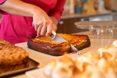 Téléchargez les photos : Confiseur méconnaissable coupe gâteau à l'aide d'un couteau tout en se tenant au comptoir de cuisine avec divers desserts sur elle - en image libre de droit