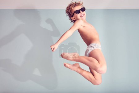 Téléchargez les photos : Vue latérale complète du corps du garçon actif en sous-vêtements et lunettes de soleil sautant haut et souriant tout en s'amusant le matin - en image libre de droit