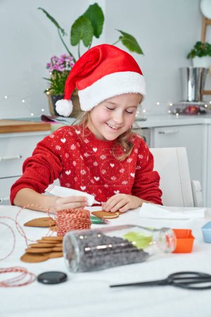 Photo for Positive girl in Santa hat applying sweet icing on tasty baked gingerbread cookie in kitchen during Christmas preparation at home - Royalty Free Image