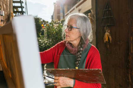 Photo for Side view of aged female artists painting on canvas in backyard on sunny day in summer - Royalty Free Image