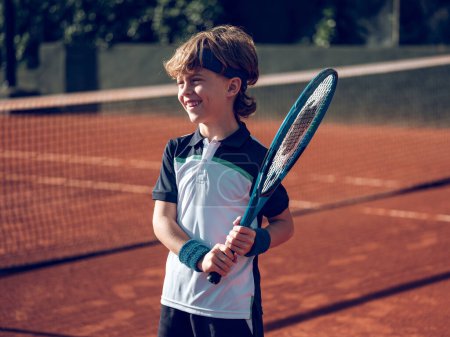 Cute smiling preteen boy in sportswear with professional tennis racket on hard court while training in sunny day