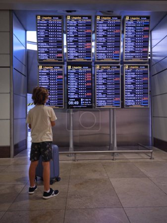 Téléchargez les photos : Vue arrière de l'enfant anonyme avec bagages regardant l'horaire d'arrivée en attendant le départ au terminal de l'aéroport - en image libre de droit