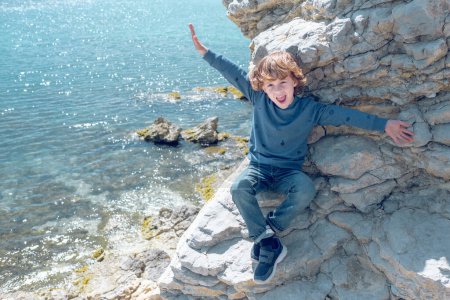 Téléchargez les photos : Du haut du corps plein de garçon positif avec les bras écartés regardant la caméra tout en étant assis sur une falaise pierreuse au-dessus de la mer - en image libre de droit