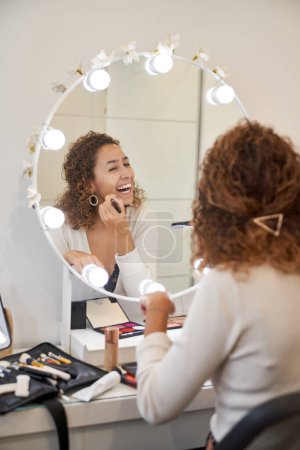 Téléchargez les photos : Vue arrière de la femme heureuse avec les cheveux bouclés appliquer fond de teint avec brosse tout en riant et en regardant miroir - en image libre de droit