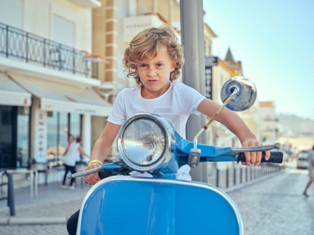 Niño con el pelo rizado haciendo cara divertida mientras monta scooter azul en la calle en Nazare en Portugal mientras disfruta de un día de verano brillante