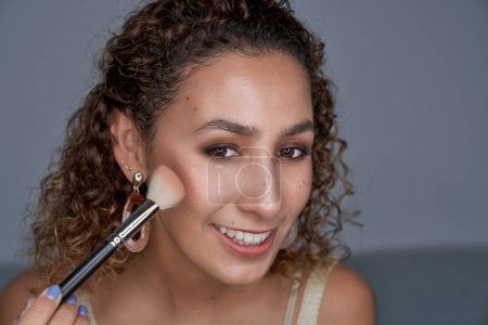 Photo for Smiling curly haired female shading blush with professional visage brush while doing makeup - Royalty Free Image