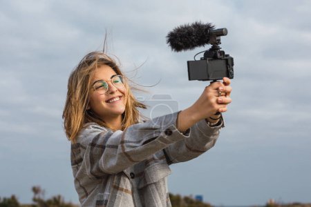 Téléchargez les photos : Joyeux jeune voyageuse aux cheveux blonds en vêtements décontractés et lunettes prenant selfie sur appareil photo numérique moderne - en image libre de droit