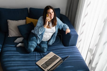 Femme enceinte freelance assis sur le canapé avec ordinateur portable dans le salon et parlant sur téléphone portable tout en travaillant à la maison