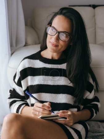 Photo for Attractive female in eyeglasses looking into distance with dreamy look and taking notes in notepad while sitting near sofa in light living room - Royalty Free Image