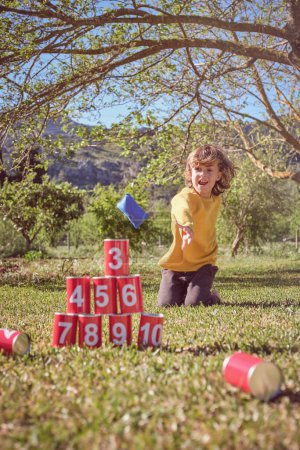Foto de Sonriente niño en suéter amarillo jugando puede Knockdown juego mientras que tirar bolsas en la pirámide de latas apiladas en el césped en el día soleado en el parque - Imagen libre de derechos