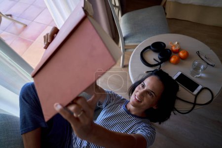 Foto de Desde arriba de libro positivo de lectura femenina hispana con los brazos levantados mientras está sentado cerca de la mesa con teléfono inteligente y auriculares en la habitación - Imagen libre de derechos