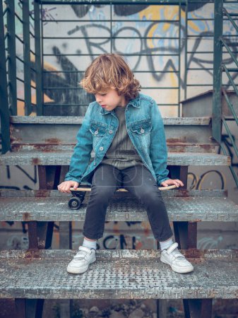 Téléchargez les photos : Corps complet de mignon garçon en vêtements décontractés assis sur le panneau de penny sur escalier minable tout en regardant vers le bas sur la rue - en image libre de droit