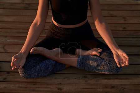 Téléchargez les photos : De dessus de la récolte anonyme pieds nus femelle en vêtements de sport assis dans la pose de lotus avec les mains mudra pendant la séance de yoga sur le chemin en bois dans la nature - en image libre de droit