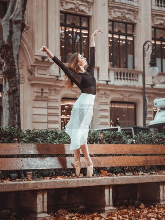 Foto de Talentosa dama con los brazos levantados mirando hacia otro lado mientras baila ballet en el banco de la calle frente al edificio en otoño - Imagen libre de derechos