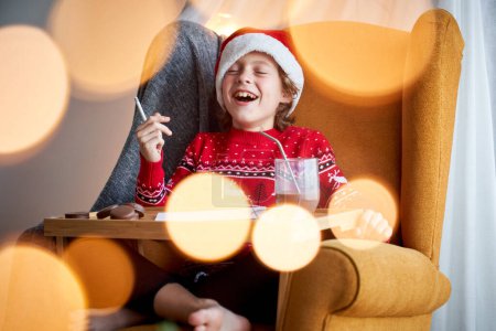 Photo for Child in Santa hat laughing in armchair while drawing on table with glass of chocolate and cookies during Christmas celebration - Royalty Free Image