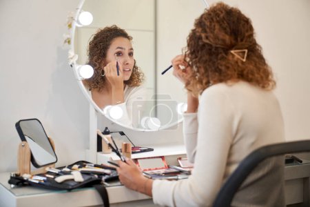 Photo for Back view of gorgeous female visagiste applying eyeshadow while sitting at table with various cosmetic products and looking at mirror - Royalty Free Image