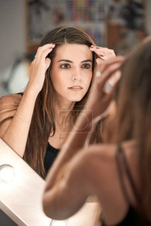 Foto de Atractiva modelo femenina tocando el pelo largo y mirando al espejo con lámparas brillantes mientras está de pie en el salón de belleza de luz moderna - Imagen libre de derechos