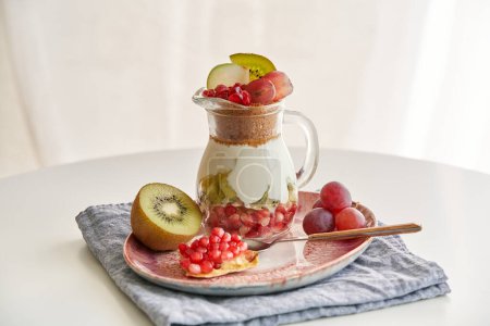 Téléchargez les photos : Grand angle de pot en verre avec délicieux dessert de yaourt et granola émietté garni de raisins et de morceaux de kiwi et de grenade grains placés sur l'assiette avec cuillère et serviette - en image libre de droit