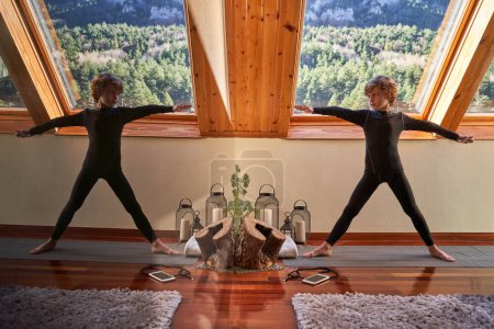 Téléchargez les photos : Corps complet d'un garçon mince en vêtements de sport noirs faisant de l'exercice tout en pratiquant le yoga sur tapis dans un appartement spacieux et en regardant le miroir - en image libre de droit