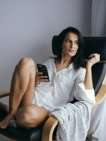 Foto de Mujer étnica descalza joven pacífica con el pelo largo y oscuro en camisa blanca mirando hacia otro lado soñando mientras usa el teléfono móvil y descansa en el sillón en casa - Imagen libre de derechos