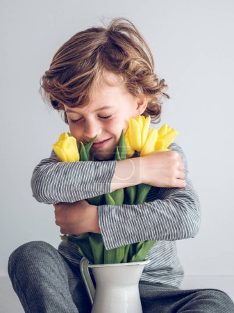 Téléchargez les photos : Garçon joyeux avec les yeux fermés embrassant bouquet de tulipes jaunes fraîches dans un vase en céramique sur fond blanc dans la pièce lumineuse - en image libre de droit