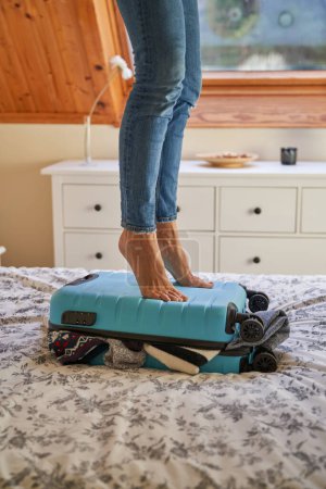 Photo for Crop unrecognizable person in jeans standing on suitcase while packing luggage on soft bed in bedroom - Royalty Free Image