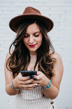 Téléchargez les photos : Cheveux longs femme attrayante dans la messagerie chapeau dans le téléphone mobile - en image libre de droit