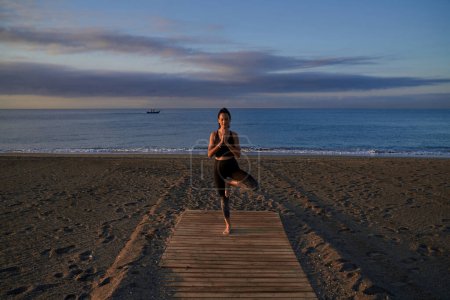Téléchargez les photos : Corps entier de femme hispanique calme pratiquant la posture Vrikshasana avec des mains namastes pendant l'entraînement de yoga sur la côte sablonneuse près de la mer - en image libre de droit