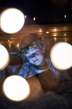 Téléchargez les photos : Adorable garçon en vêtements de nuit jouer au jeu sur tablette tout en étant assis dans la chambre sombre près de guirlande et bokeh sur la nuit - en image libre de droit