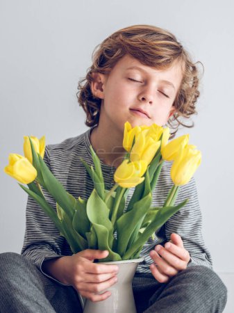 Téléchargez les photos : Adorable enfant sentant le bouquet de fleurs de tulipes jaunes dans un vase et profitant d'un parfum frais avec les yeux fermés sur fond blanc - en image libre de droit