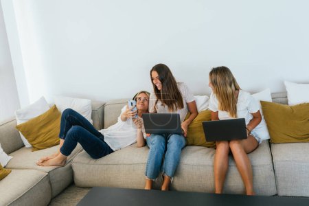 Photo for Woman in headphones showing video on cellphone to cheerful female friends with laptops while sitting on couch in living room - Royalty Free Image