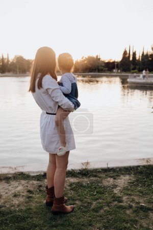 Téléchargez les photos : Vue arrière de la jeune femme tenant la petite fille debout sur le rivage près de l'eau ondulante tout en passant du temps dans le parc en soirée - en image libre de droit