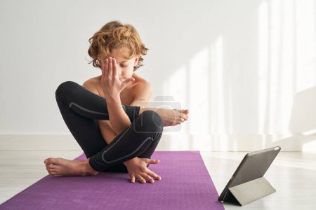 Téléchargez les photos : Garçon aux cheveux blonds posant d'embryon dans l'utérus tout en faisant du yoga sur le sol contre la tablette dans un appartement léger - en image libre de droit