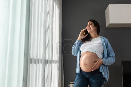 Téléchargez les photos : Sourire femme enceinte toucher le ventre tout en se tenant debout dans la chambre à la maison et parler téléphone mobile - en image libre de droit