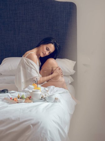 Téléchargez les photos : Vue latérale de la femelle sereine en vêtements de nuit en soie se détendre sur le lit près du plateau avec petit déjeuner savoureux frais et profiter de la matinée dans un hôtel de luxe - en image libre de droit