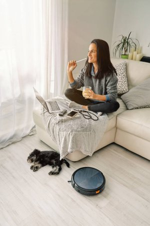 Téléchargez les photos : Vue latérale de la femme aux cheveux bruns assise avec verre de boisson sur canapé avec ordinateur portable ouvert et smartphone dans la chambre avec chien endormi et aspirateur robotisé - en image libre de droit