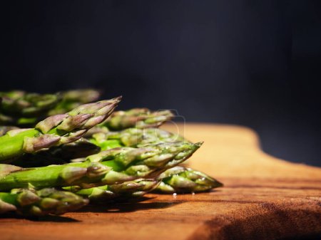 Photo for Pile of fresh uncooked green healthy asparagus placed on wooden cutting board against dark background in kitchen during cooking preparation - Royalty Free Image