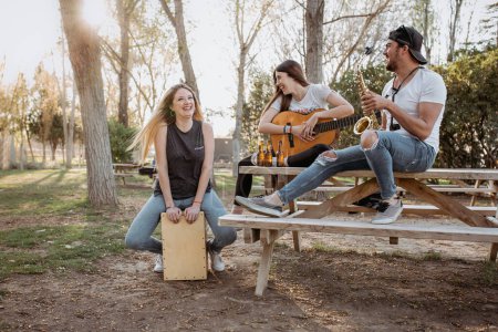 Foto de Joven hombre y mujeres con instrumentos musicales sentados cerca de la mesa y riendo en un día soleado en el parque - Imagen libre de derechos