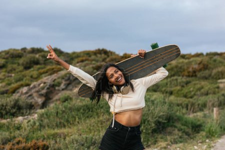 Photo for Young smiling ethnic cheerful female standing with longboard behind head and headphones on neck looking at camera showing peace gesture - Royalty Free Image