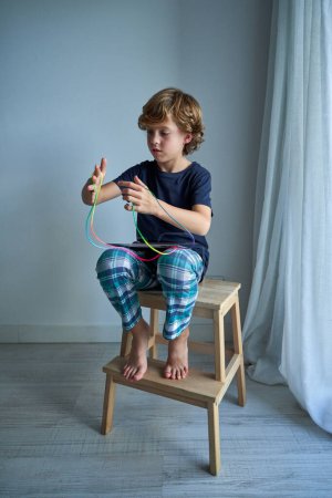 Foto de Cuerpo completo de niño descalzo enfocado en ropa casual jugando juego de cuerdas mientras está sentado con la tableta en el taburete de madera en la sala de estar en casa - Imagen libre de derechos