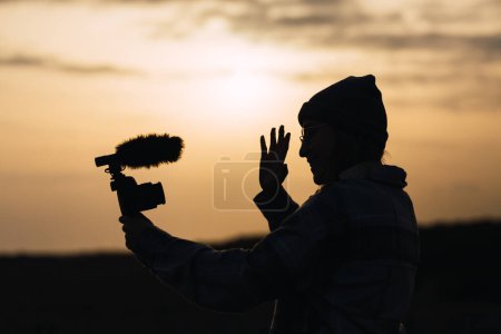 Foto de Silueta de vista lateral de una joven blogger irreconocible con sombrero sonriendo y mostrando la mano ondeando mientras filma video en cámara digital contra el cielo del atardecer en el campo - Imagen libre de derechos