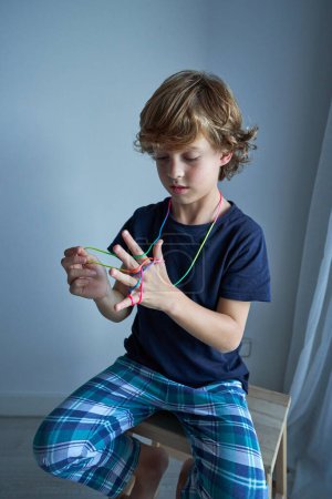 Photo for Portrait of focused child sitting on wooden stool while playing with rainbow finger thread and learning to make string figures at home - Royalty Free Image