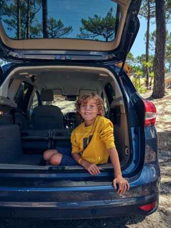 Foto de Longitud completa del niño sentado en el maletero del automóvil y mirando a la cámara durante las vacaciones de verano en el tiempo soleado en los bosques - Imagen libre de derechos