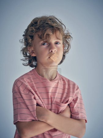 Foto de Lindo niño descontento en camisa roja mirando a la cámara con la boca sellada contra el fondo gris en el estudio y cruzando brazos - Imagen libre de derechos