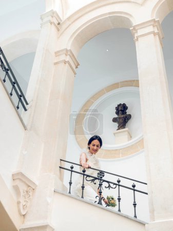 Téléchargez les photos : D'en bas de jeune femme nouvellement mariée marchant en bas sur l'escalier à l'intérieur classique du couloir avec des éléments voûtés et des colonnes et des sculptures et regardant vers le bas - en image libre de droit