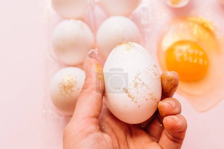 From above unrecognizable person holding festive glittered Easter egg during preparation for holidays on pink background