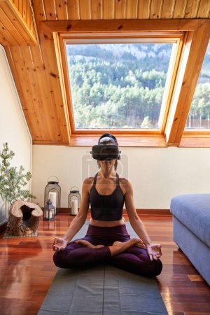 Photo for Full body of barefoot woman wearing VR goggles sitting on yoga mat and meditating in Lotus pose with crossed legs and doing mudra gesture with hands in mansard bedroom - Royalty Free Image