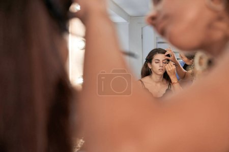 Foto de Cosecha anónima borrosa maestro con cepillo aplicar sombra de ojos en el párpado de cliente femenino sentado contra el espejo en el salón de belleza de la luz - Imagen libre de derechos