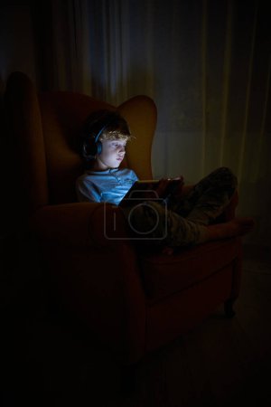 Photo for Side view of serious boy in wireless headphones playing videogame on modern tablet while sitting in dark room at home - Royalty Free Image