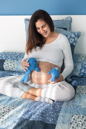 Foto de Alto ángulo de encantadora hembra embarazada sentada en una cama suave y jugando con juguetes de peluche - Imagen libre de derechos
